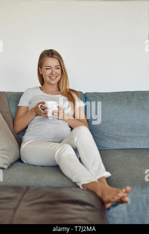 Jolie jeune femme enceinte pieds nus sur un canapé relaxant de boire une tasse de thé chaud et souriant joyeusement à l'appareil photo Banque D'Images