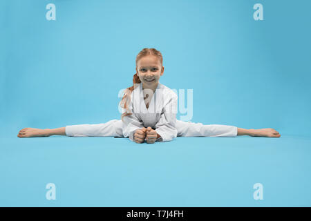 Vue avant du happy pretty girl sitting on cross et de la ficelle à la caméra à l'arrière-plan. isolé sur fond bleu Smiling child wearing kimono et s'étend au cours de la formation en studio. Concept de karaté. Banque D'Images
