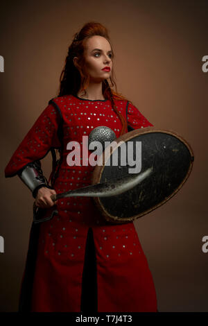 Beau, brave femme portant en rouge tunique moyenâgeuse holding bouclier et dague. Sérieux, jeune femme avec des cheveux de gingembre à l'écart, posant en studio sur fond uni. Banque D'Images