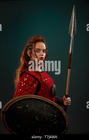 Brave femme guerrière portant en rouge costume médiéval looking at camera, tenant le bouclier et grande lance. Belle, belle femme aux longs cheveux gingembre posing in studio avec fond plein. Banque D'Images