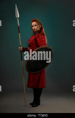 Brave femme guerrière avec l'expression du visage grave tenue longue lance et bouclier protecteur. Belle femme avec cheveux gingembre portant en rouge tunique moyenâgeuse, looking at camera, posant debout. Banque D'Images