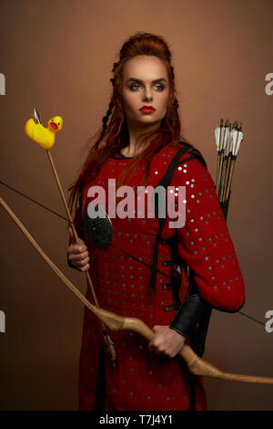 Sérieux, femme courageuse, guerrier médiéval tenant arc et flèche avec canard en caoutchouc. Superbe, jolie femme portant en tunique rouge à l'écart, posant en studio. Banque D'Images