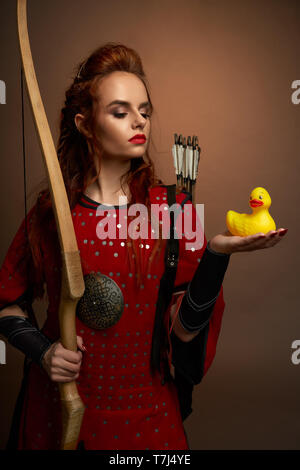 Belle guerrière portant à l'époque médiévale tunique rouge, jaune petits canards en caoutchouc sur le côté, le regardant. Modèle avec sérieux et lèvres rouges cheveux gingembre posing holding, arc. Banque D'Images