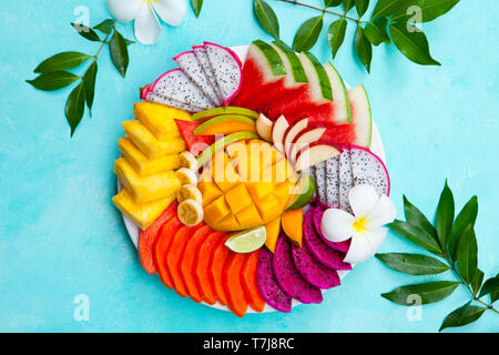 Assortiment de fruits tropicaux sur une plaque avec des fleurs de frangipanier. Fond bleu. Vue d'en haut. Copier l'espace. Banque D'Images