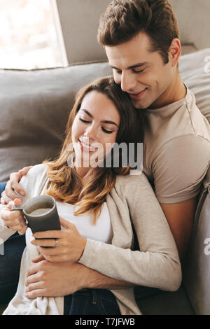 Smiling young couple relaxing sur un canapé à la maison, holding cup Banque D'Images