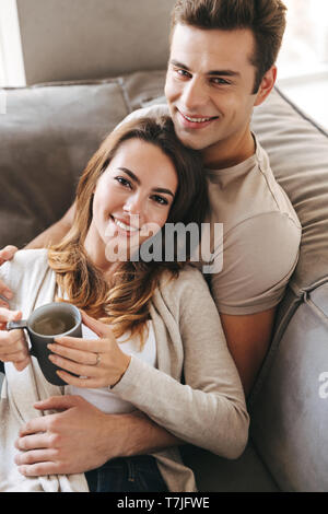 Smiling young couple relaxing sur un canapé à la maison, holding cup Banque D'Images