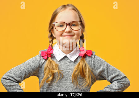 Portrait of a cute little baby girl avec des lunettes sur un fond jaune. La lycéenne fait mains dans ses hanches et se penche sur l'appareil photo. Le concep Banque D'Images