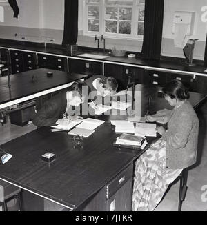 Années 1960, historique, dans une classe de l'école secondaire vide, une femme professeur de mathématiques assis à un bureau de superviser deux élèves de sexe masculin au même bureau d'étude supplémentaires faisant travailler après la classe, dans le sud de Londres, Angleterre, Royaume-Uni. Banque D'Images