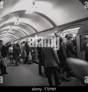 Années 1960, historiques, le métro de Londres, un train de tube à une plate-forme, les gens monter et descendre du train, Londres, Angleterre, Royaume-Uni. L'orgins du métro de Londres se trouvent dans le métro, le premier passager de métro Chemin de fer qui a ouvert ses portes en 1863. En bois éclairé au gaz voitures étaient tractés par des locomotives à vapeur. Banque D'Images