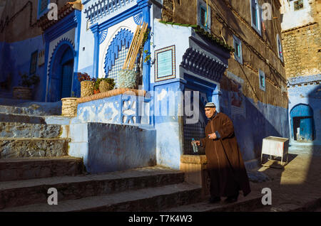 Chefchaouen, Maroc : un homme portant une djellaba remplit une bouteille d'eau d'une fontaine dans le bleu à la chaux ruelles de la médina, vieille ville. Banque D'Images