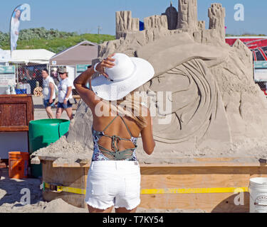 Une femme blonde bronzée en short blanc de derrière une sculpture de sable vues tout en maintenant son chapeau blanc contre le vent. 2019 Texas Sandfest. Banque D'Images