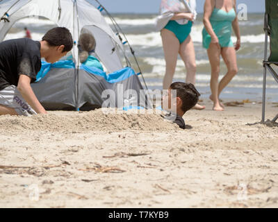 Un garçon est enterré dans le sable sur une journée ensoleillée à la plage à Port Aransas, Texas USA. Banque D'Images