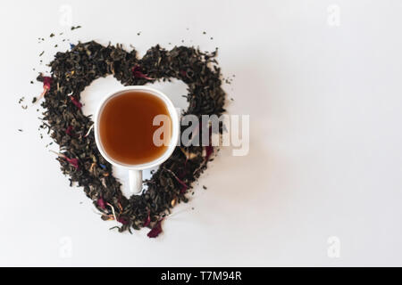 Forme de coeur fait avec un mélange d'une variété de feuilles de thé séchées et une tasse de thé noir sur fond blanc Banque D'Images