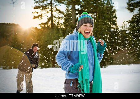 Laughing young woman wearing hat un tricot de se faire frapper par une boule de neige de derrière tandis que dehors dans la neige avec des amis. Banque D'Images