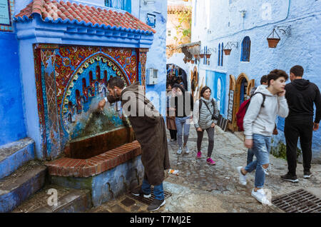 Chefchaouen, Maroc : Un homme boit de l'eau d'une fontaine dans le bleu à la chaux medina vieille ville. Banque D'Images