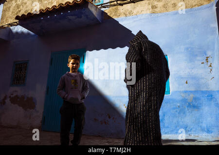 Chefchaouen, Maroc : un homme portant une trational djellaba et un stand de l'enfant par un mur lavé dans la médina vieille ville. Banque D'Images