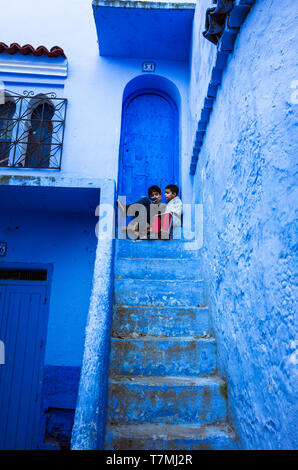 Chefchaouen, Maroc : deux enfants sont assis sur le dessus d'un escalier bleu à la chaux dans la médina, vieille ville. Banque D'Images