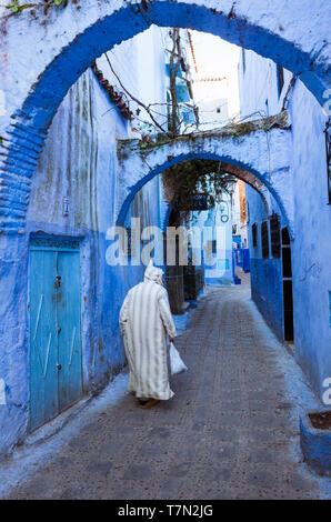 Chefchaouen, Maroc : un homme portant une djellaba traditionnelle promenades dans le bleu à la chaux ruelles de la médina, vieille ville. Banque D'Images