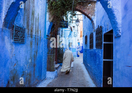 Chefchaouen, Maroc : un homme portant une djellaba traditionnelle promenades dans le bleu à la chaux ruelles de la médina, vieille ville. Banque D'Images