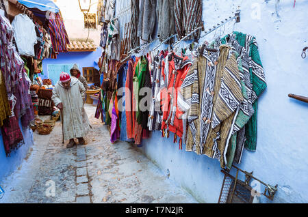 Chefchaouen, Maroc : un homme portant une djellaba passe devant un magasin de vêtements dans le bleu à la chaux medina vieille ville. Banque D'Images