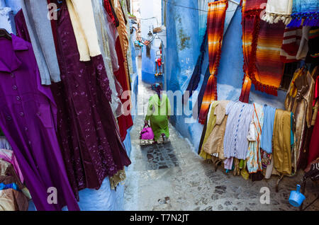 Chefchaouen, Maroc : une femme passe devant un magasin de vêtements dans le bleu à la chaux medina vieille ville. Banque D'Images