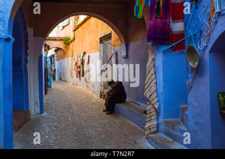 Chefchaouen, Maroc : Un homme est assis sous une arche dans le bleu à la chaux ruelles de la médina, vieille ville. Banque D'Images