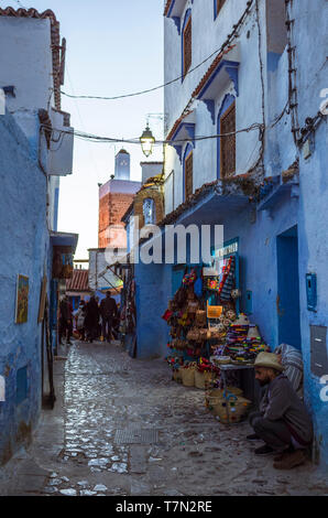 Chefchaouen, Maroc : deux hommes assis à la tombée de l'extérieur d'une boutique dans les ruelles de la médina bleu à la chaux vieille ville. Minaret illuminé en arrière-plan. Banque D'Images