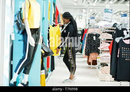 Afican femmes américaines en survêtements et lunettes de shopping au centre commercial de sport contre les étagères. Elle a choisi un t-shirt jaune. Sport store thème. Banque D'Images