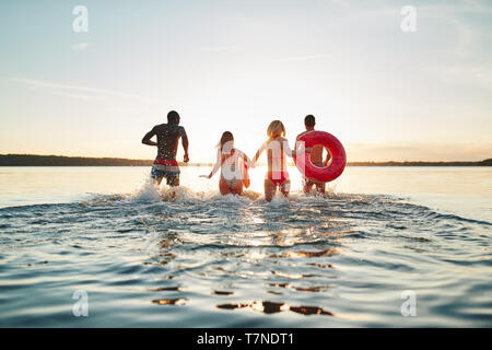 Coquille d'un groupe diversifié de jeunes amis en maillot de bain d'exécution dans un lac au coucher du soleil les projections d'eau et faire des vagues Banque D'Images