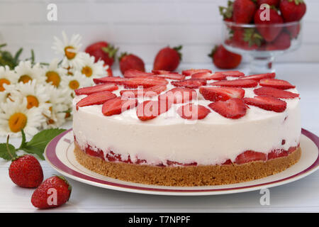 Un gâteau aux fraises sans cuisson, avec une base de biscuits, est placé à l'horizontale sur une plaque sur un fond blanc avec un bouquet de marguerites dans Banque D'Images