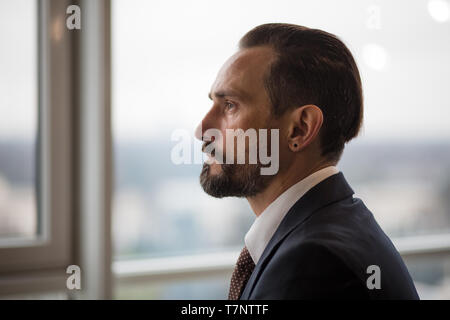 Elegant businessman dans le bureau en regardant par la fenêtre. Il a l'air très sérieux. Il est pensif. Il porte un costume noir a une moustache et b Banque D'Images