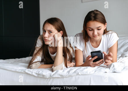 Deux filles mignon teengae portant sur le lit, tenant un téléphone mobile à la maison Banque D'Images