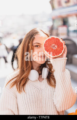 Portrait de jeune fille à la mode belle attrayante avec des écouteurs est titulaire de la moitié d'agrumes en part, couvrant son œil Banque D'Images