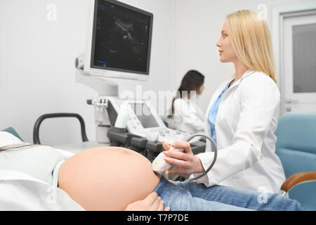 Voir du côté de la femme médecin sérieux en uniforme blanc faisant de femme enceinte de diagnostic à ultrasons et à la recherche à l'écran dans une clinique. Femme avec gros ventre couché sur coach. Concept de la médecine. Banque D'Images