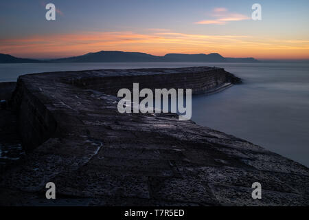 La célèbre Lyme Regis, Cobb, qui serpente vers la mer avec le soleil se lever sur les collines au loin, la longue exposition à lisser l'océan. Banque D'Images