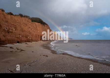 Un arc-en-ciel touche le sol sur une plage de Sidmouth dans le Devon, en Angleterre, avec les falaises rouges et ciel bleu Banque D'Images