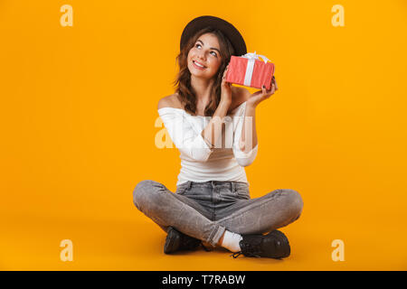 Portrait d'une jeune femme portant une chemise blanche et un chapeau assis sur fond jaune isolé, holding gift box Banque D'Images