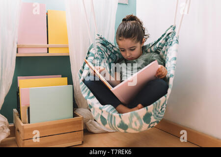 Ambiance petite fille lire un livre dans son hamac près de la fenêtre, drôle belle enfant s'amusant dans sa chambre d'enfant Banque D'Images