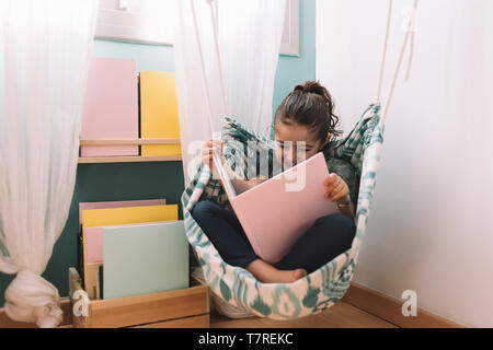 Petite fille de rire en lisant un livre dans son hamac près de la fenêtre, drôle belle enfant s'amusant dans sa chambre d'enfant Banque D'Images