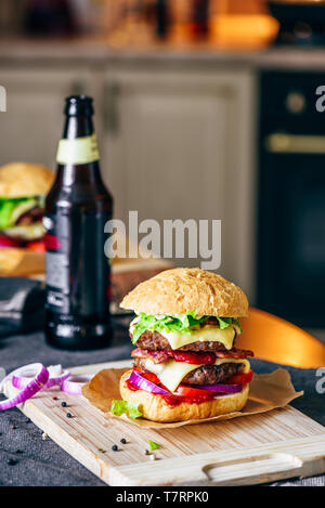 Cheeseburger avec deux galettes de boeuf, fromage cheddar, bacon, laitue Iceberg, des tranches de tomate et d'oignon rouge. Bouteille de bière et des ingrédients sur tabl Banque D'Images