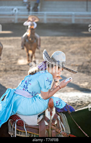 Saray Franco avec le légendaire Franco famille de champions Charro, regarde son téléphone portable en attendant que son cas au cours d'une session pratique rodéo mexicain de Jalisco dans la ville de Highlands Capilla de Guadalupe, au Mexique. Les participantes à l'Escaramuza Charreada traditionnels sont appelés et effectuer la precision des manifestations équestres amazone et vêtue en robe Adelita. Banque D'Images