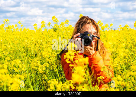 Jeune femme photographe avec un reflex numérique, portant une veste orange, prend une photo dans un champ de colza, campagne, l'Europe de l'Est, Roumanie - Copie Banque D'Images