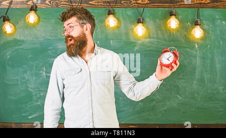 Régime de l'annexe et concept. Enseignant dans les lunettes est titulaire d'un réveil. Hipster barbu est titulaire d'réveil, tableau sur toile de fond, l'espace de copie. Homme à barbe à crier face à argumenter l'expression. Banque D'Images