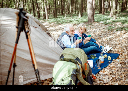 Senior couple sitting ensemble au camping avec tente et sacs à dos, profiter de la nature et de boire du café dans la forêt Banque D'Images