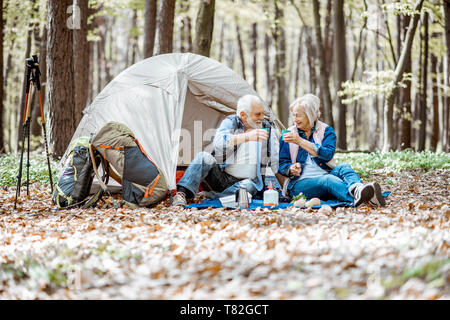 Senior couple sitting ensemble au camping avec tente et sacs à dos, profiter de la nature et de boire du café dans la forêt Banque D'Images