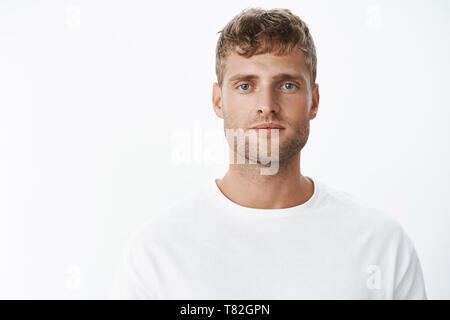 Taille plan sur beau calm blonde blue-eyed guy avec en soie blanc T-shirt looking at camera avec l'expression du visage insouciant détendue posant plus de Banque D'Images