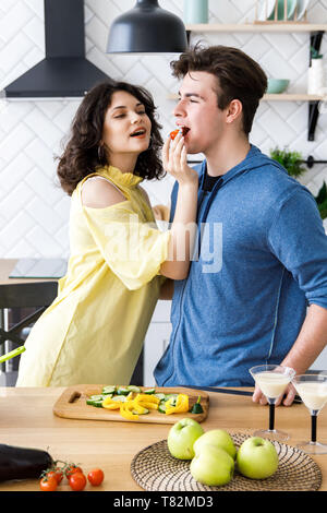 Young cute smiling couple cooking ensemble en cuisine à la maison. Cute girl nourrit son petit ami avec petites tomates rouges Banque D'Images