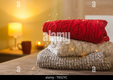 Pile de pulls sur un lit décoré de lumières. Petite lampe, bougie et tasse sur la table en bois en arrière-plan. Concept confortable chaleureux. Banque D'Images