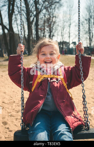 Peu souriant heureux fille se balançant dans un parc aux beaux jours du printemps. Enfant looking at camera wearing red jacket Banque D'Images