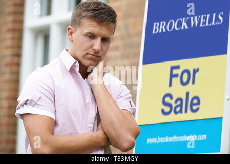 Jeune homme forcé de vendre la maison à travers des problèmes financiers à l'extérieur debout à côté de For Sale Sign Banque D'Images